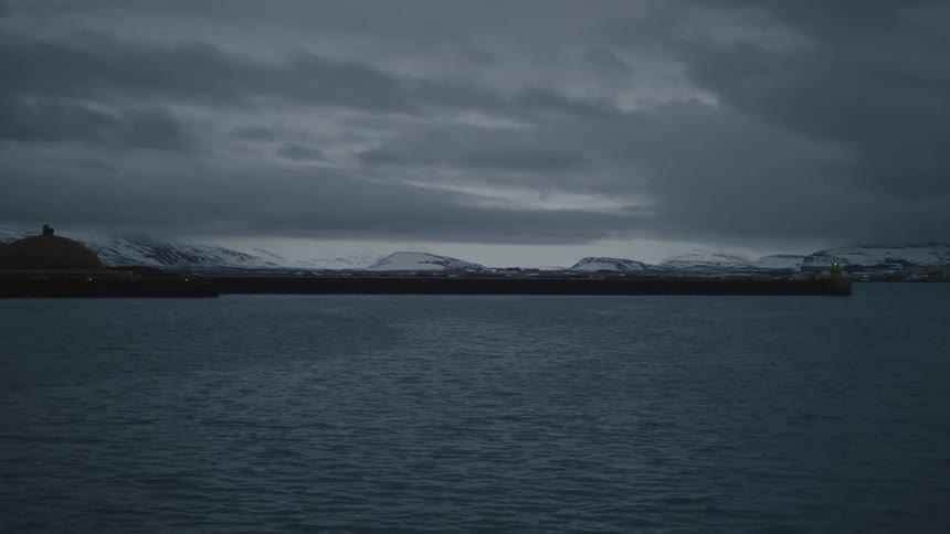Lights blink on the waterfront under an overcast sky with snowy mountains in the distance
