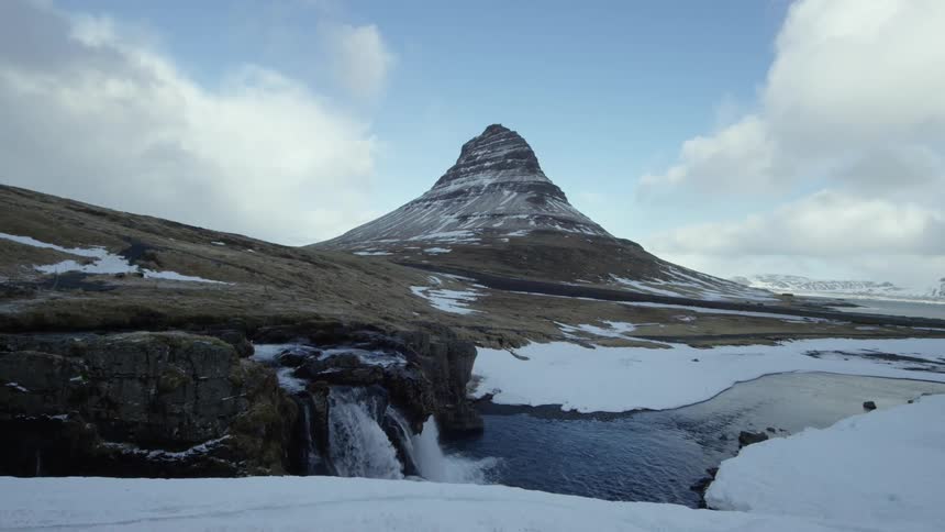 Snowcapped conical mountain stands out in the background under a partly cloudy sky and in the foreground a waterfall flows into a partially frozen river