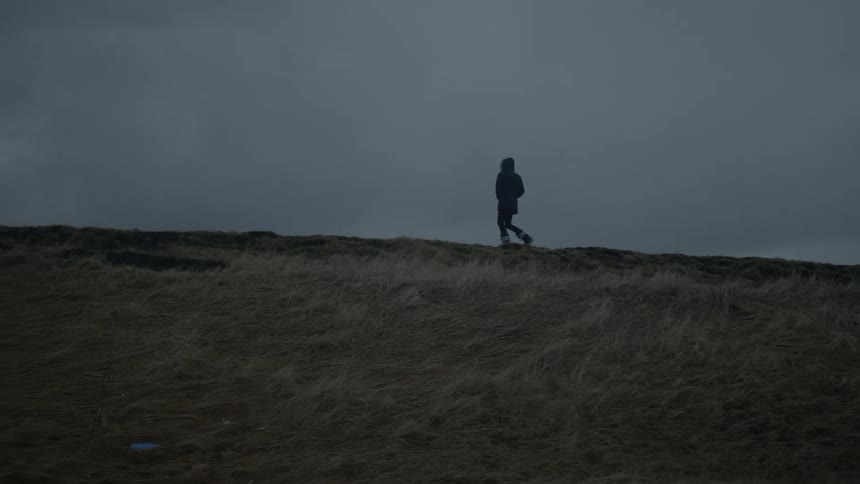 Person walking on a grassy hill against a dimly lit cloudy sky