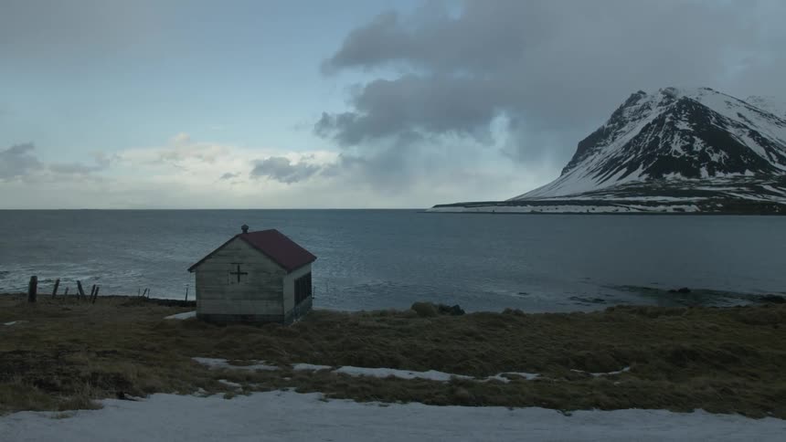 Small wooden house on the edge of a lake with snowcovered mountains in the background