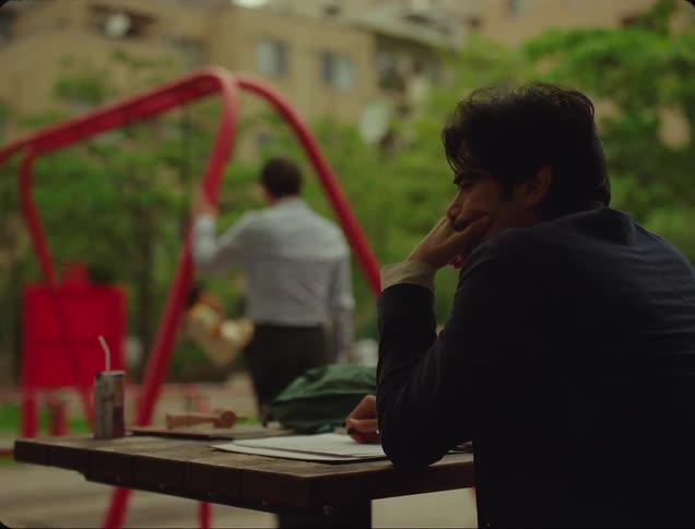 Man sitting at a table thinking and in the background a father watching his child playing in a square