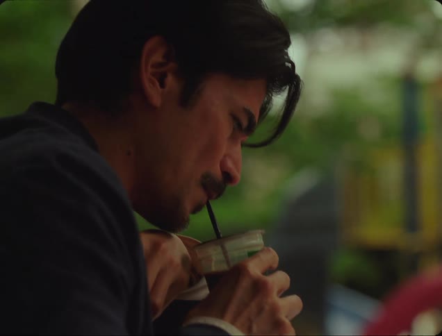Man sitting at a table outdoors while thinking takes a sip of a coffee drink through a straw