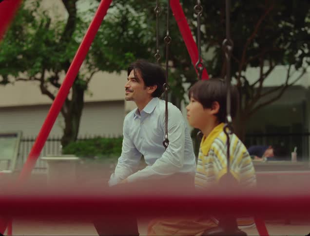 A father sits on a swing next to his son and smiles at him