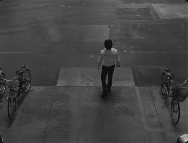A person in a white shirt and dark pants walks on a gray pavement flanked by parked bicycles