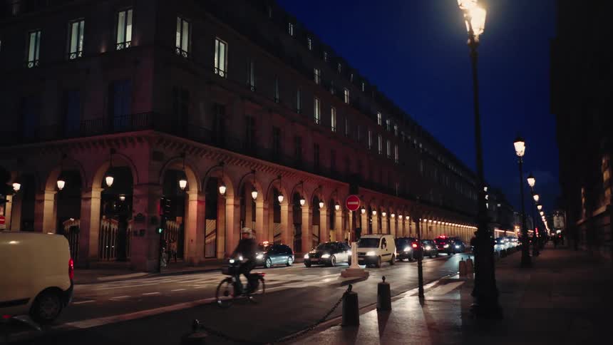 Two bicyclists ride down an urban street at night while traffic is stopped at a light in Paris