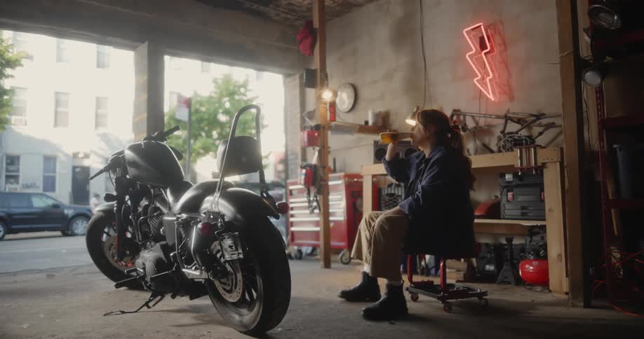 A female mechanic sits in a repair shop and drinks a beer