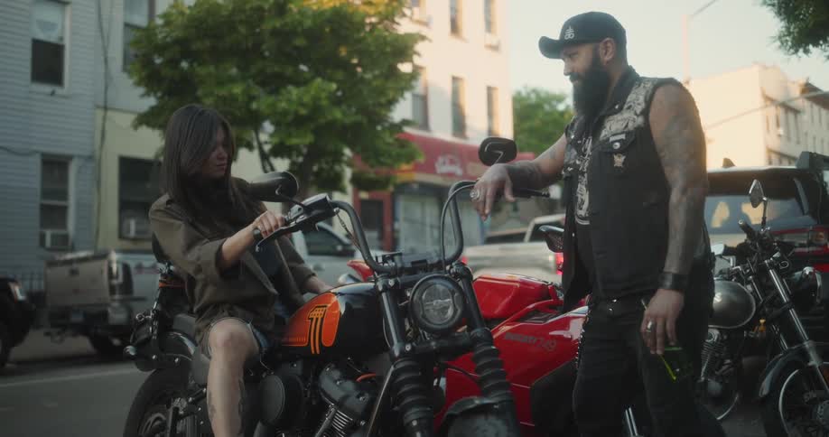 A young woman sitting on a bikers motorcycle to test it as he watches her