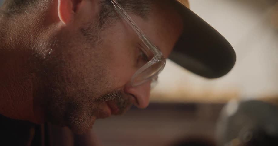 A man wearing glasses and a hat looks down and concentrates on his task