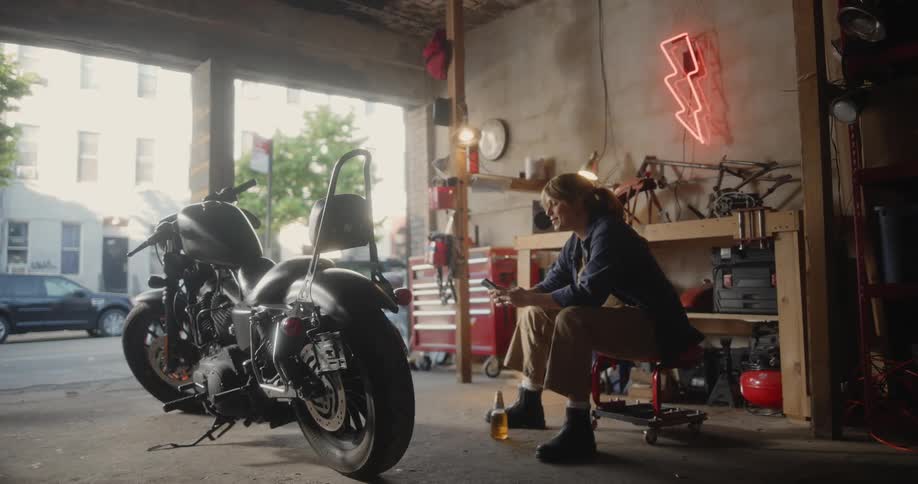 A mechanic sits on a stool in a garage next to a classic motorcycle texting on her phone