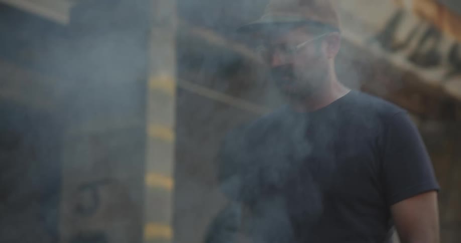 A man wearing glasses cooks food on a grill outdoors as people talk nearby