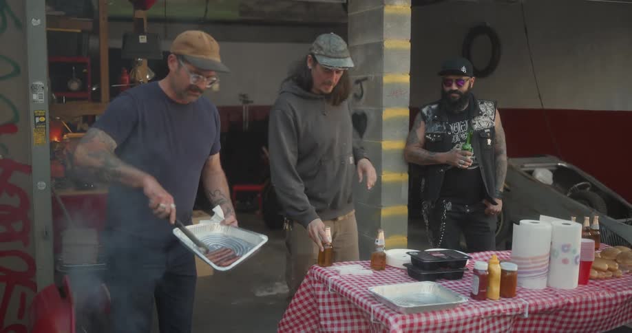 A group of men gathered around a table with food and drinks enjoying a casual outdoor barbecue