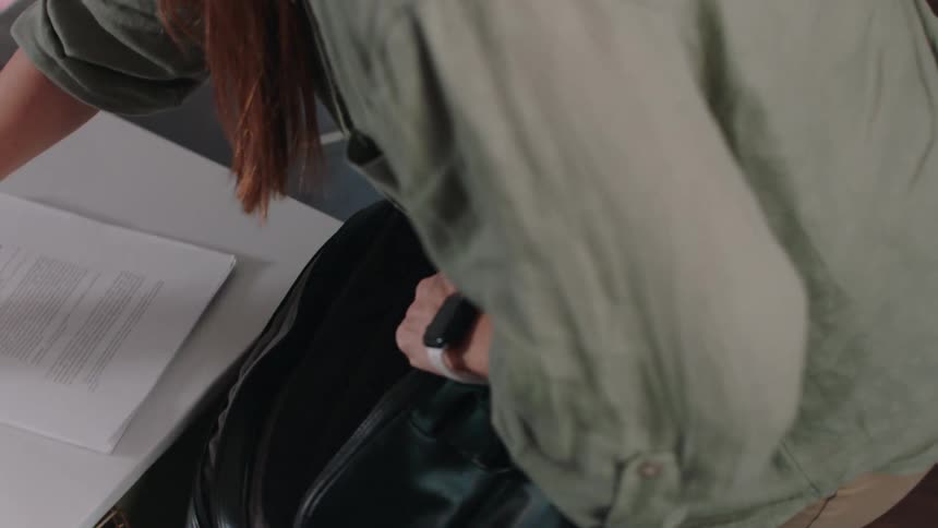 Female student puts her textbooks and other school items into a book bag while standing in the living room