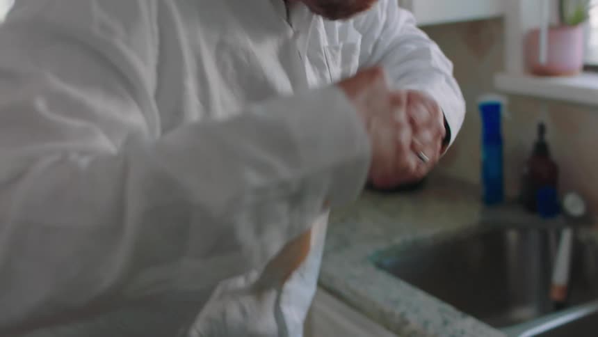 A man walks in his kitchen as he tries to drink coffee then spits it out because its too hot staining his shirt then uses a paper wipe to clean his mouth