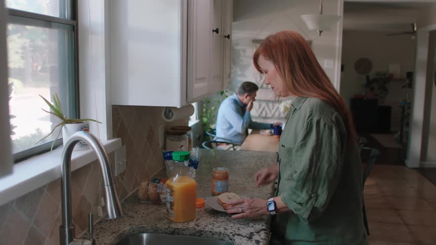 Woman putting peanut butter on a slice of bread and in the background her husband sitting at the table