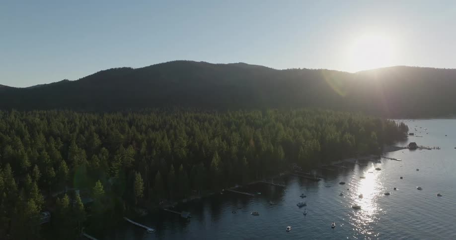 A bay of water with some boats and on the shore there is a pine forest and in the background there is a mountain range with the sun coming out from behind them