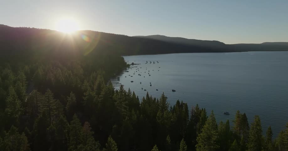 Aerial landscape of a lake surrounded by mountains and conifers with the sun on the horizon