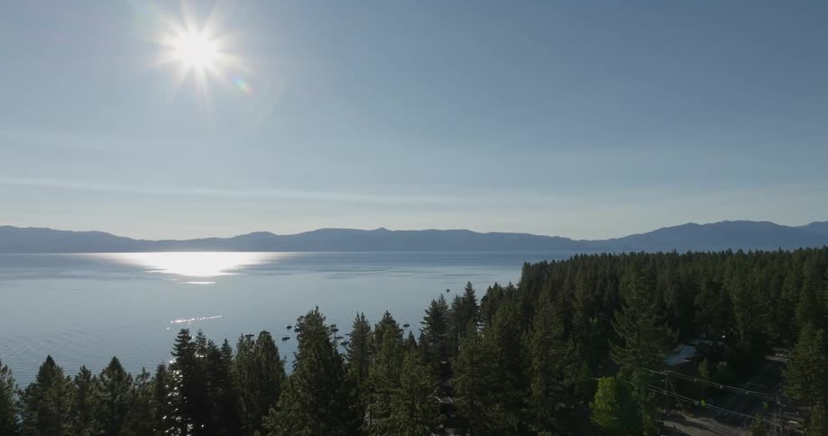 Aerial landscape of conifers in the foreground next to a highway and in the background a vast lake surrounded by mountains with the sun highlighted in a blue sky
