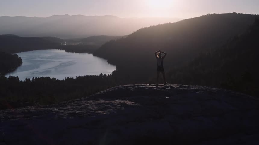 Woman on a mountain peak raising her arms with a view of a lake landscape and a sun on the horizon