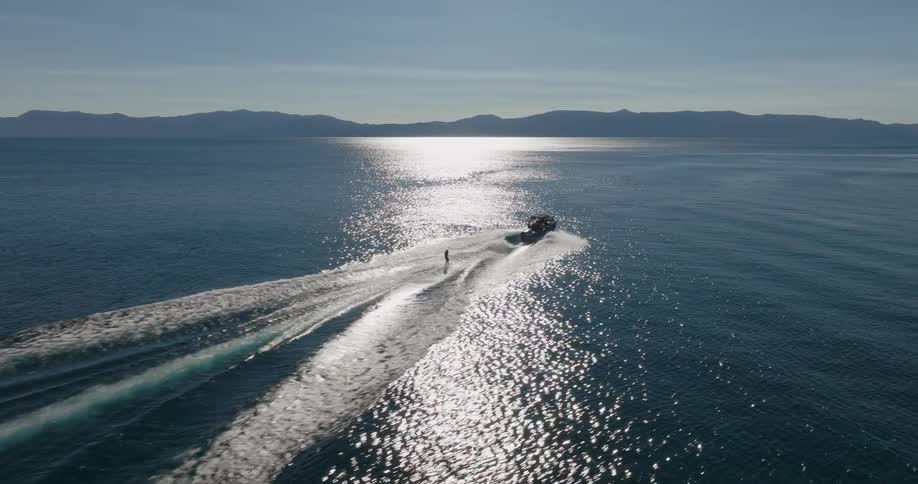 A speedboat on a large body of water creating a wake as it moves behind a person is wakeboarding on the glistening surface