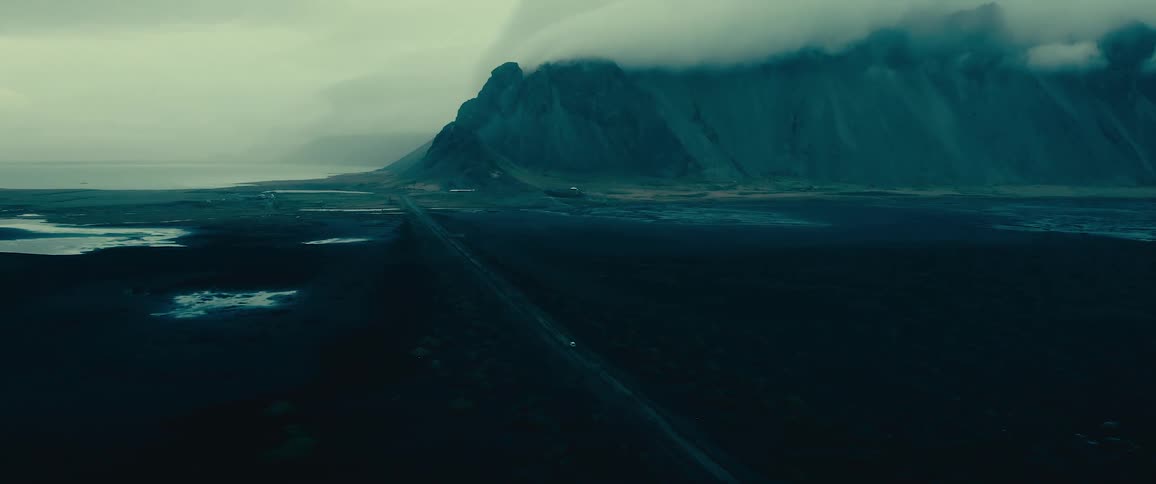 Aerial view of a vehicle driving through a vast flat landscape with a straight road leading toward a large mountain range enveloped in clouds