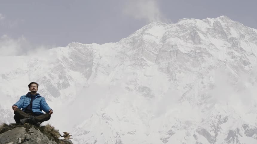 An individual sits on a rock legs crossed facing away from a vast mountain range covered in snow