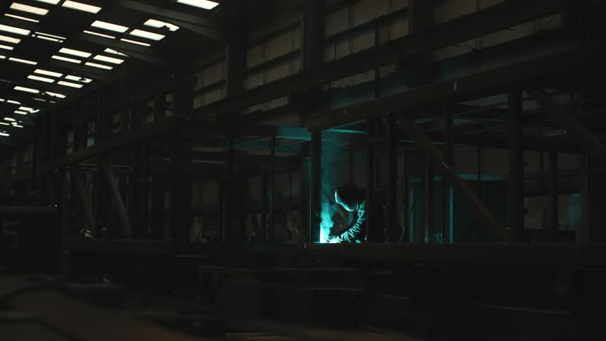 Person wearing protective helmet working with a welding tool that emits smoke glow and sparks inside a factory