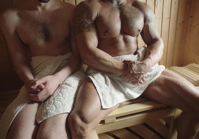 A strong muscular man sits uncomfortably close to another man in a sauna