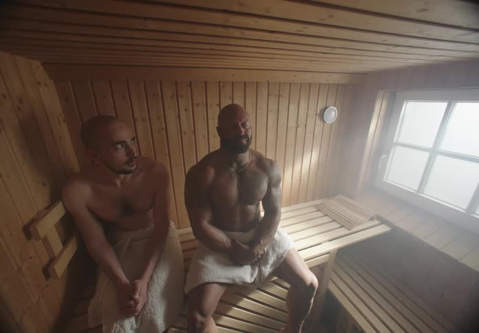Two men sitting in a wooden sauna one with a muscular build and the other with a shaved head both wrapped in towels