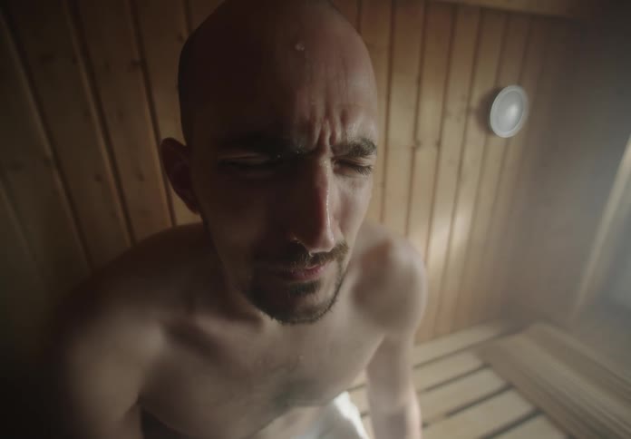 A man sits on a bench in a wooden sauna room surrounded by steam with sweat pouring down his face