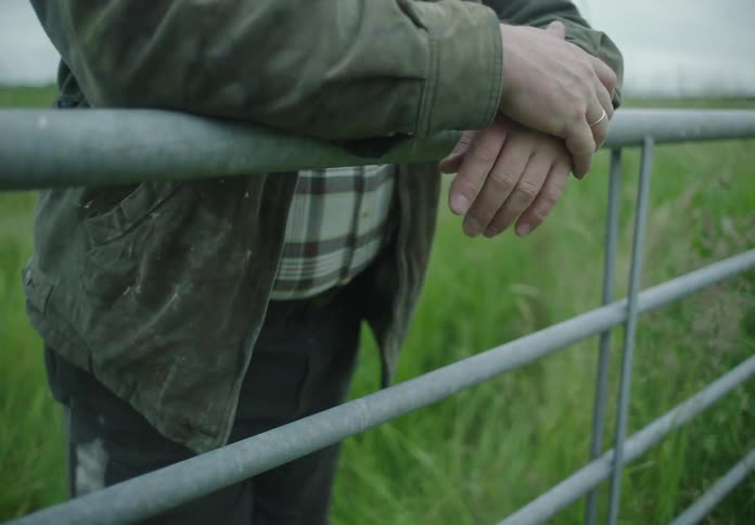 A white male walks through a grassy field and leans against a metal fence