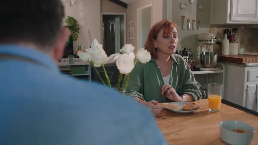 Mother sitting at the table looking at her wristwatch and her son runs past the kitchen sitting at the table with his parents with a bowl
