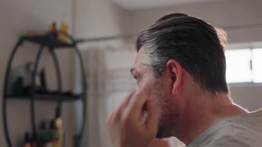 A man rubbing his face and hair in front of the bathroom mirror