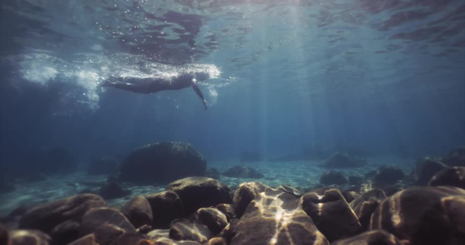 Underwater footage of a woman swimming in a clear mountain lake with sunlight filtering through the surface illuminating a bed of rounded rocks