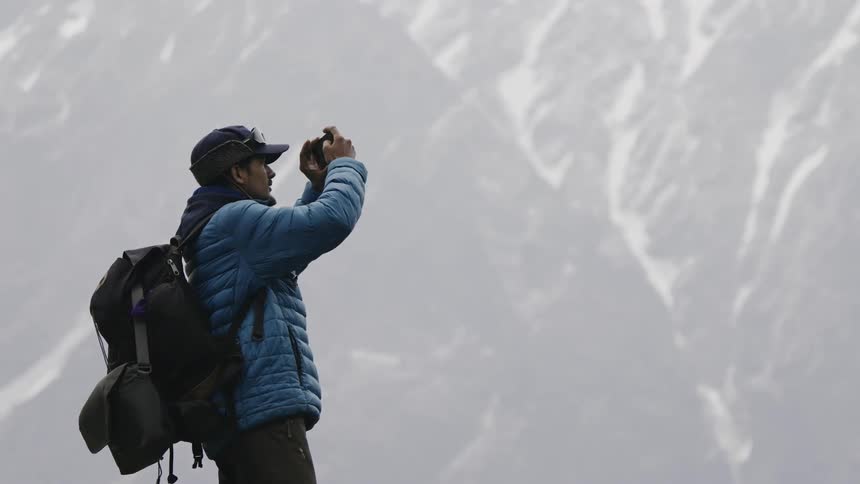 A person in a blue jacket and backpack taking a panoramic photo with a smartphone of a snowy mountain range