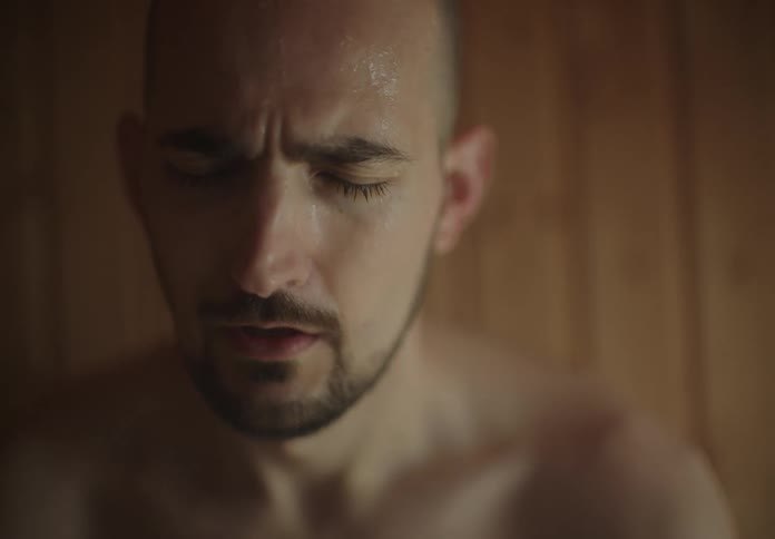 A man sits in a wooden sauna room surrounded by steam with sweat pouring down his face