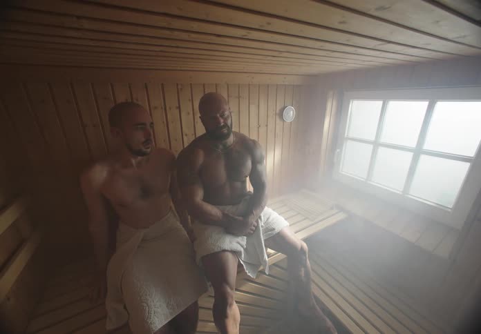 Uncomfortable man next to a muscular man as they both sit side by side in a sauna