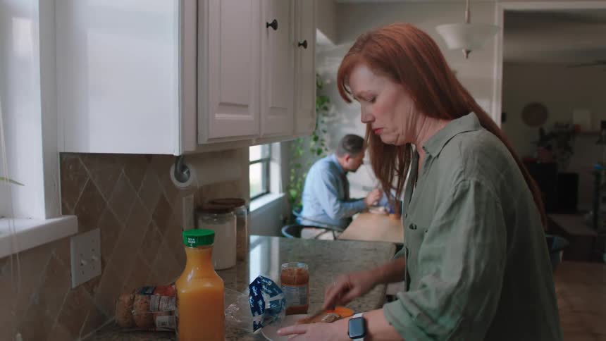 A woman preparing breakfast in the kitchen at home her husband is already eating in the background