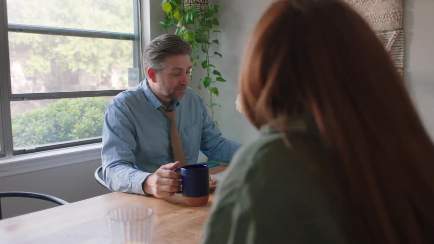Man sitting down drinking a cup and a woman sitting together at the table and talking