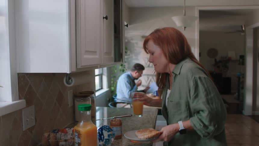 Woman placing a glass of juice and a plate with two breads sitting at the table next to her husband