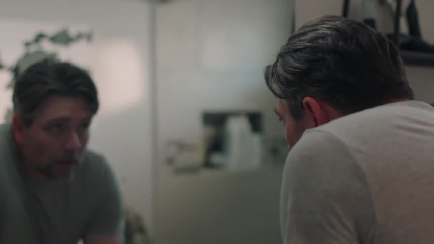A man bends over a sink and looks at himself in a bathroom mirror