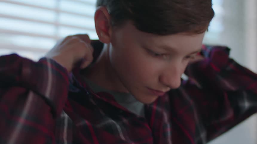 A teenage boy is buttoning his shirt while standing indoors