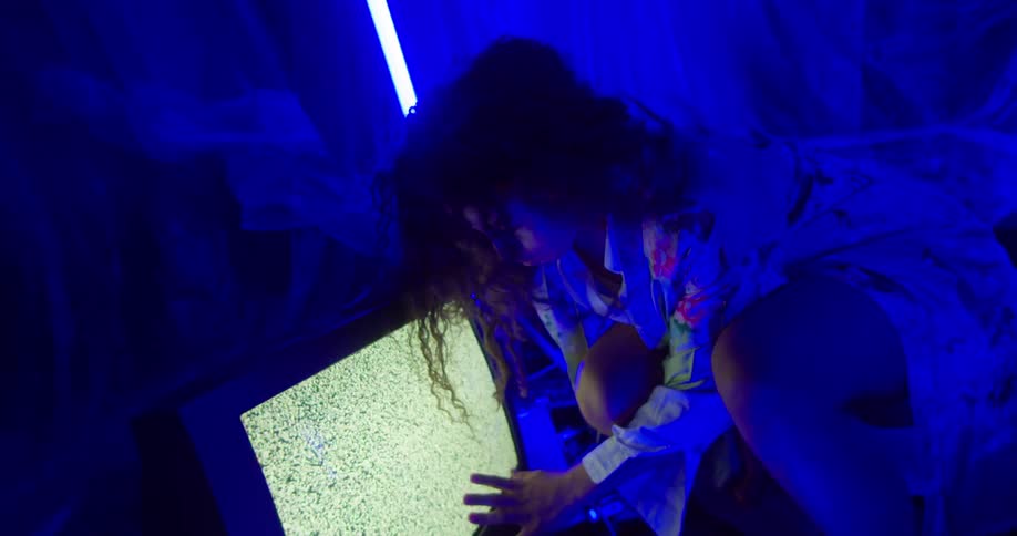 A woman with curly hair and a flowery dress leans over a vintage television with static on the screen