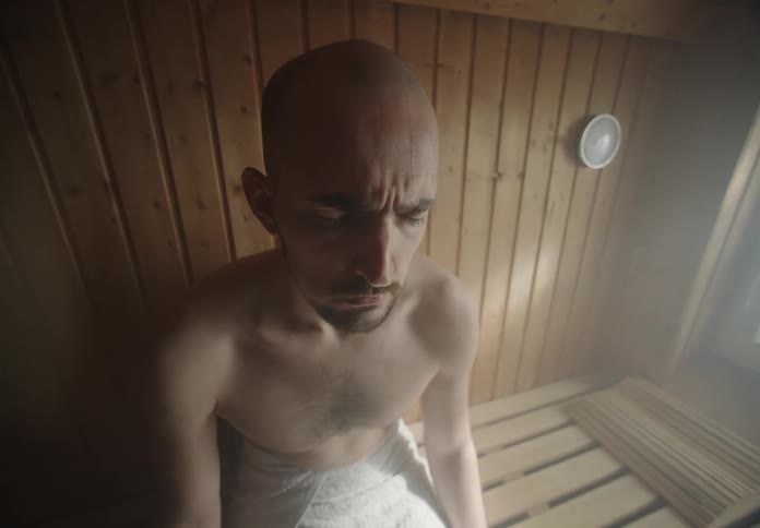 A man sits on a bench in a wooden sauna room surrounded by steam with sweat pouring down his face