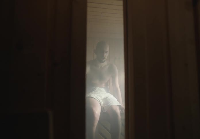 A man sitting in a sauna partially obscured by steam and wooden panels