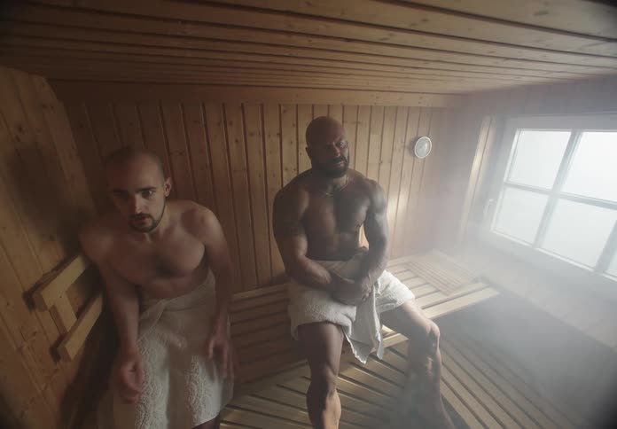 Man walking away from another muscular man standing next to him inside a sauna