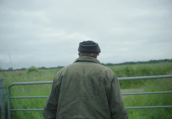 Back view of a man walking towards a fence in a field with tall grass