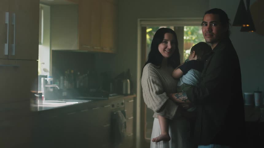 A man and a woman standing in a kitchen both holding a baby together as they all turn and smile to the camera