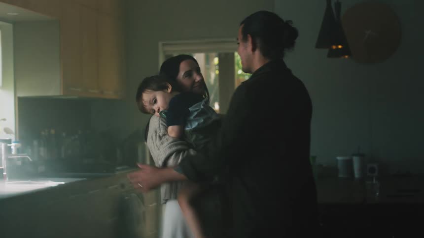 Man with jacket and long hair holds womans hands picking up toddler son in the kitchen