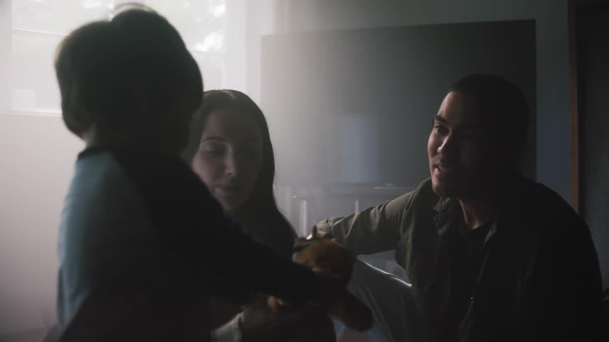 A man and woman sit in a dark room with their young son and the mom puts a toy on the boys stuffed animal and he shakes it