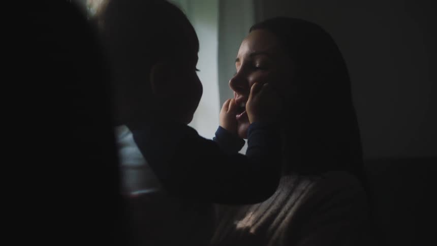 A woman holding a toddler boy in her arms as he squishes her face with his hands and she squeezes him making him laugh